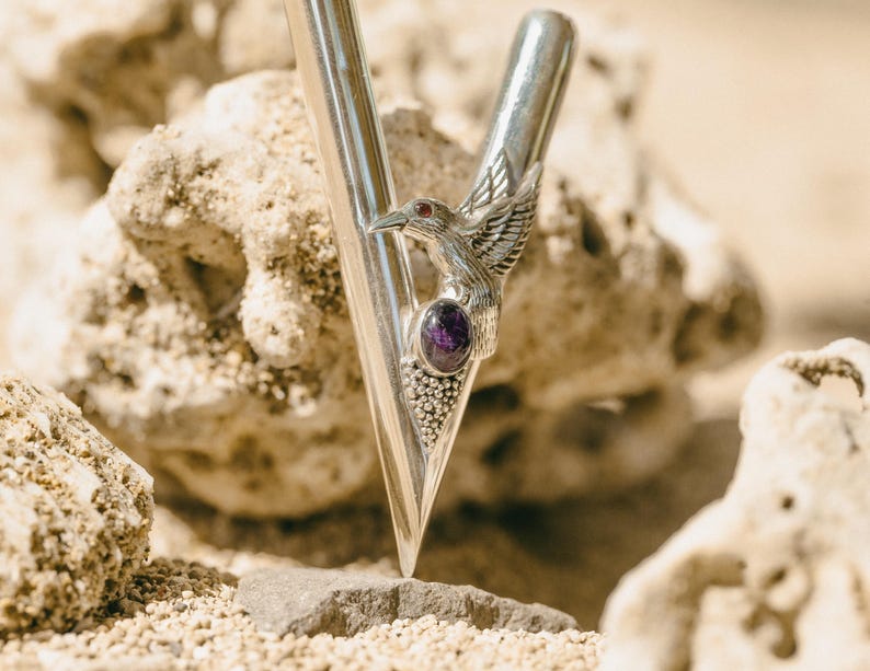 Puede incluir: Una horquilla de metal plateado con un detalle de p&aacute;jaro y piedra preciosa p&uacute;rpura. La horquilla tiene forma de V con un extremo puntiagudo y una parte superior redondeada. El p&aacute;jaro tiene las alas extendidas y est&aacute; posado sobre la piedra preciosa.