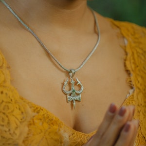 Puede incluir: Un collar de plata con un colgante de tridente detallado con una piedra preciosa verde. El collar lo lleva una persona con una blusa de encaje amarilla.