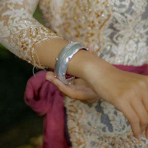 Könnte beinhalten: Ein silbernes Armband in Federform mit einer strukturierten Oberfläche. Das Armband wird am Handgelenk getragen.