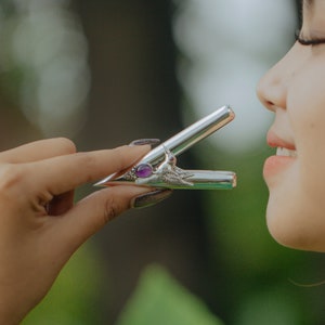Puede incluir: Un atomizador de perfume plateado con un dise&ntilde;o decorativo y una piedra preciosa p&uacute;rpura. El atomizador se sostiene cerca de la nariz. El dise&ntilde;o incluye un motivo de p&aacute;jaro o ala.