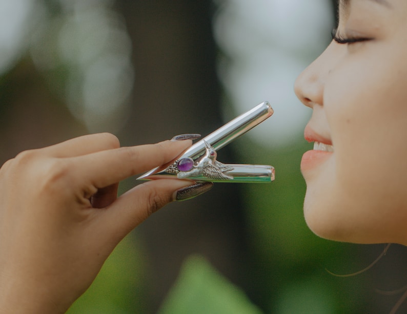 Puede incluir: Un atomizador de perfume plateado con un dise&ntilde;o decorativo y una piedra preciosa p&uacute;rpura. El atomizador se sostiene cerca de la nariz. El dise&ntilde;o incluye un motivo de p&aacute;jaro o ala.