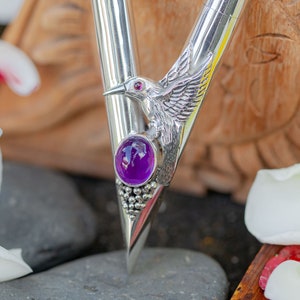 Puede incluir: Un bol&iacute;grafo plateado con un dise&ntilde;o de colibr&iacute;. El bol&iacute;grafo presenta una piedra preciosa p&uacute;rpura y un ojo de piedra preciosa roja en el colibr&iacute;. El fondo est&aacute; borroso, con p&eacute;talos de flores blancas.