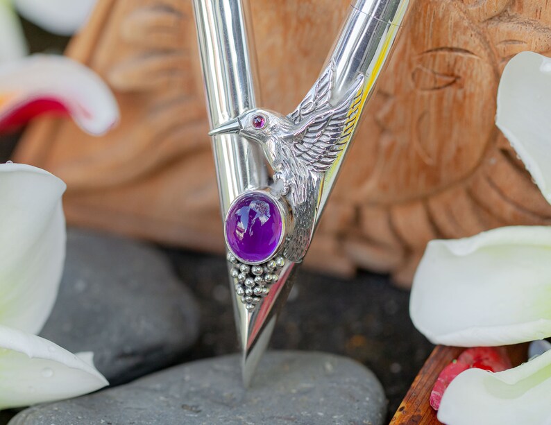 Puede incluir: Un bol&iacute;grafo plateado con un dise&ntilde;o de colibr&iacute;. El bol&iacute;grafo presenta una piedra preciosa p&uacute;rpura y un ojo de piedra preciosa roja en el colibr&iacute;. El fondo est&aacute; borroso, con p&eacute;talos de flores blancas.