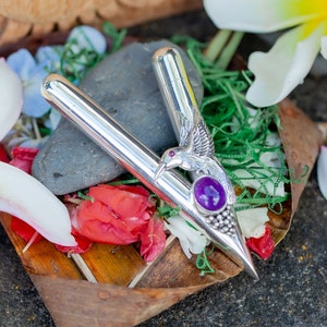 Puede incluir: Un objeto plateado en forma de V con un dise&ntilde;o de colibr&iacute; y una piedra preciosa p&uacute;rpura. El objeto est&aacute; rodeado de flores, vegetaci&oacute;n y una piedra. El colibr&iacute; tiene alas detalladas y un pico largo. La piedra es ovalada.