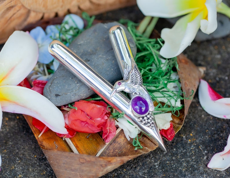 Puede incluir: Un objeto plateado en forma de V con un dise&ntilde;o de colibr&iacute; y una piedra preciosa p&uacute;rpura. El objeto est&aacute; rodeado de flores, vegetaci&oacute;n y una piedra. El colibr&iacute; tiene alas detalladas y un pico largo. La piedra es ovalada.