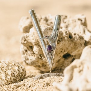 Puede incluir: Un colgante de metal plateado en forma de V con un dise&ntilde;o de colibr&iacute; y una piedra preciosa p&uacute;rpura. El colgante se encuentra sobre un fondo de rocas y arena beige, creando una est&eacute;tica natural y elegante.