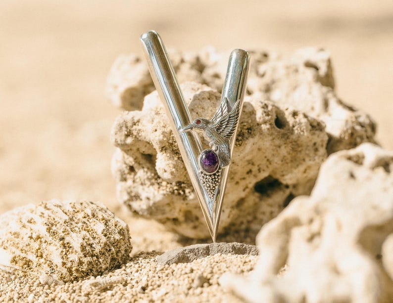 Puede incluir: Un colgante de metal plateado en forma de V con un dise&ntilde;o de colibr&iacute; y una piedra preciosa p&uacute;rpura. El colgante se encuentra sobre un fondo de rocas y arena beige, creando una est&eacute;tica natural y elegante.