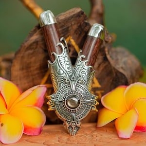May include: A silver and wood decorative pendant with intricate carvings and a gemstone in the center. The pendant is surrounded by two yellow flowers with pink edges.
