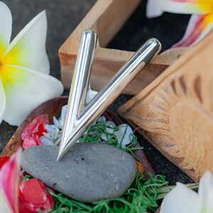 Puede incluir: Una herramienta de masaje plateada en forma de V, apoyada sobre una piedra gris. La herramienta est&aacute; rodeada de flores coloridas, hierba verde y elementos decorativos de madera. Una flor blanca y amarilla est&aacute; en el fondo.
