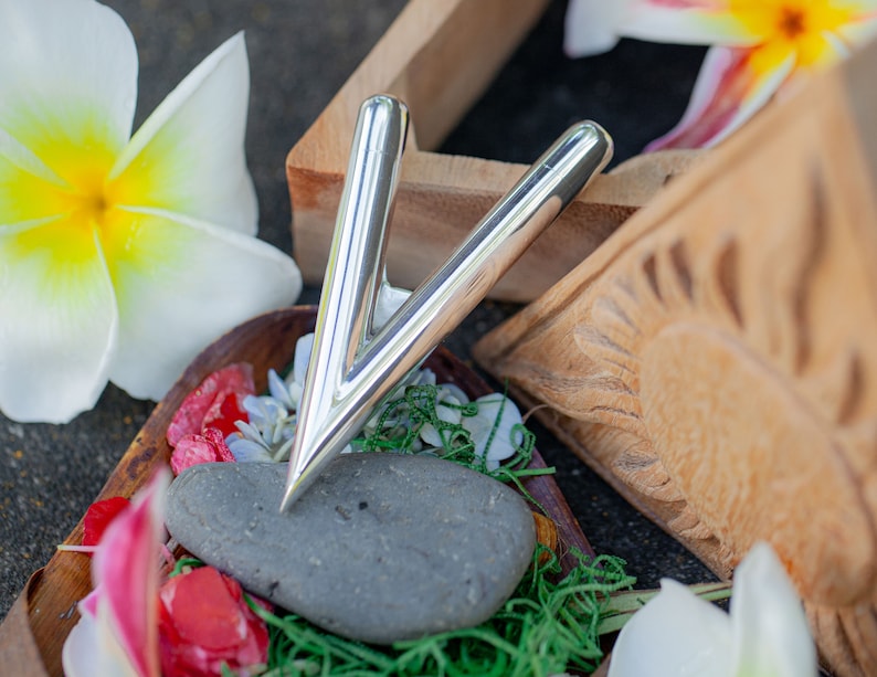 Puede incluir: Una herramienta de masaje plateada en forma de V, apoyada sobre una piedra gris. La herramienta est&aacute; rodeada de flores coloridas, hierba verde y elementos decorativos de madera. Una flor blanca y amarilla est&aacute; en el fondo.