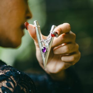 Puede incluir: Accesorio met&aacute;lico plateado con dise&ntilde;o en forma de V, con una gema p&uacute;rpura y un emblema de p&aacute;jaro. El accesorio se sostiene en una mano con esmalte de u&ntilde;as rojo, sobre un fondo verde borroso.