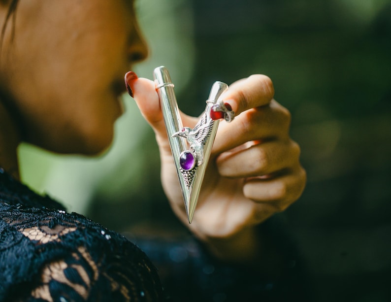 Puede incluir: Accesorio met&aacute;lico plateado con dise&ntilde;o en forma de V, con una gema p&uacute;rpura y un emblema de p&aacute;jaro. El accesorio se sostiene en una mano con esmalte de u&ntilde;as rojo, sobre un fondo verde borroso.