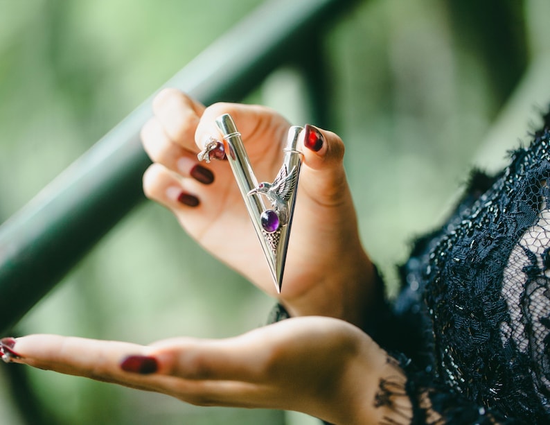 Puede incluir: Una pinza de corbata plateada con un dise&ntilde;o triangular, con una piedra preciosa p&uacute;rpura y un emblema de &aacute;guila. La pinza de corbata se sostiene en una mano con esmalte de u&ntilde;as rojo oscuro, sobre un fondo verde borroso.
