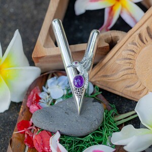 Puede incluir: Un bol&iacute;grafo plateado con un dise&ntilde;o en forma de V, con un colibr&iacute; y una piedra preciosa p&uacute;rpura. El bol&iacute;grafo se presenta con flores y un elemento de madera tallada, creando una presentaci&oacute;n decorativa y art&iacute;stica.