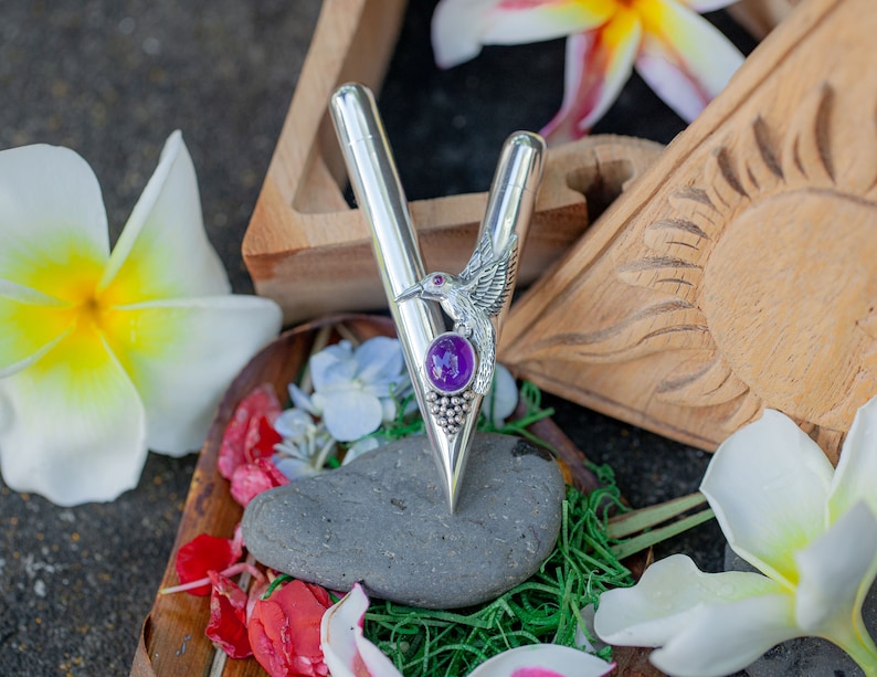 Puede incluir: Un bol&iacute;grafo plateado con un dise&ntilde;o en forma de V, con un colibr&iacute; y una piedra preciosa p&uacute;rpura. El bol&iacute;grafo se presenta con flores y un elemento de madera tallada, creando una presentaci&oacute;n decorativa y art&iacute;stica.