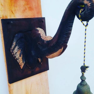 May include: A wooden elephant head sculpture with a bell hanging from its trunk. The sculpture is mounted on a wooden plaque with a brown and orange painted surface.
