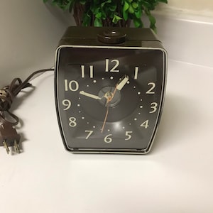 Puede incluir: Un reloj despertador vintage marrón con una esfera blanca y agujas negras. El reloj tiene una esfera redonda con números negros y un borde blanco. El reloj está enchufado con un cable.