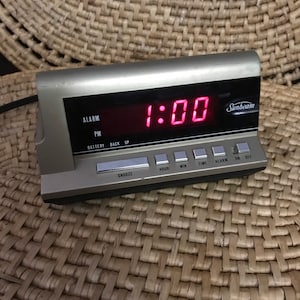 May include: A silver Sunbeam digital alarm clock with a red LED display showing 1:00. The clock has buttons for alarm, snooze, hour, minute, time, and on/off.