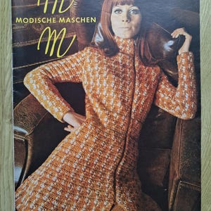Puede incluir: Una portada de revista vintage con una mujer con un abrigo naranja y blanco estampado con cremallera. El título de la revista, "Modische Maschen", se muestra en letras doradas. La mujer está sentada en un sillón de cuero marrón.
