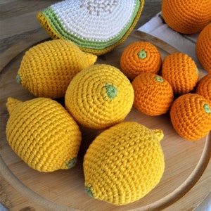 Könnte beinhalten: Ein Set gehäkelter Früchte, darunter Zitronen und Orangen, auf einem Holzbrett. Die Früchte sind aus gelbem und orangefarbenem Garn gefertigt, und die Zitronen haben grüne Blätter.