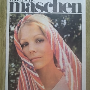 Może przedstawiać: Wydanie archiwalnego czasopisma Modische Maschen z 1976 roku, opublikowane przez Verlag für die Frau w Lipsku. Na okładce znajduje się osoba w wielokolorowej, dzianinowej chuście w paski w odcieniach pomarańczu, różu i bieli na ciemnym tle.