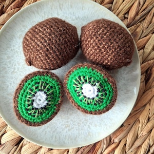 Può includere: Un piatto bianco contiene quattro kiwi all'uncinetto. Due kiwi interi sono marroni e due sono tagliati, rivelando dettagli verdi, bianchi e neri. Il piatto è su una superficie intrecciata marrone chiaro.