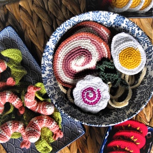 Può includere: Una ciotola in ceramica blu e bianca piena di cibi all'uncinetto, tra cui un vortice rosa e bianco, un uovo giallo e verdure a foglia verde. La ciotola è su una superficie di vimini intrecciato.