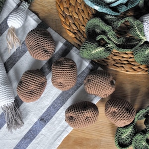 Peut inclure: Un ensemble de légumes en crochet, comprenant des pommes de terre, des poireaux et un navet, sur un fond de tissu rayé. Les légumes sont faits de fil brun, blanc et vert.