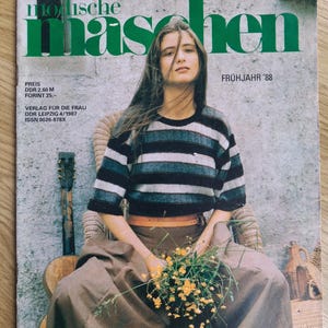 Può includere: Copertina di rivista con una donna in maglione a righe e gonna lunga, con fiori gialli. Il titolo della rivista è "modische maschen" in verde, con testo che indica una pubblicazione del 1988.