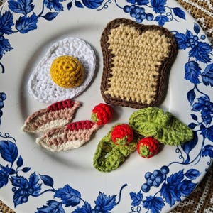 Puede incluir: Un juego de desayuno de ganchillo sobre un plato blanco con diseños florales azules. El juego incluye un huevo frito, una rebanada de pan tostado, tiras de tocino y tomates con lechuga. Los artículos son hechos a mano.