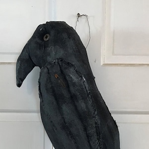 May include: Handmade black fabric crow decoration with a button eye and safety pin detail. The crow has a long, curved beak and a stitched body. A wire loop is attached for hanging. A rustic, gothic-themed home decor item.