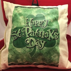 May include: Cream-colored pillow with a green shamrock design and the text "Happy St. Patrick's Day." The shamrock has a textured, distressed look. The pillow is displayed on a red surface.