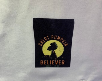 May include: White fabric tote bag with a black patch featuring a cartoon dog in front of a yellow moon with the text "Great Pumpkin Believer Since 1966".