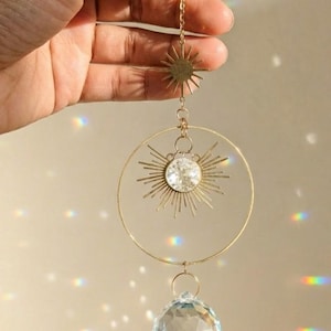 Könnte beinhalten: Ein goldfarbener Sonnenfänger mit Sonnenstrahlendesign, einer klaren Kristallkugel und einem großen kreisförmigen Rahmen. Der Sonnenfänger wird von einer zarten Kette gehalten. Die Kristallkugel ist facettiert und reflektiert das Licht. Er wird von einer Hand gehalten.