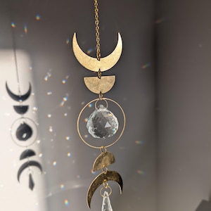 Op de afbeelding: Een hemels thema hangende decoratie met goudkleurige halve manen, halve manen en een kristallen bol. Het ontwerp bevat een ketting en een druppelvormige kristallen hanger. Het zonlicht creëert een regenboogeffect op de muur.