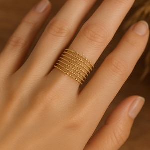 Könnte beinhalten: Ein goldfarbener Ring mit einem mehrschichtigen Design. Der Ring hat sieben Bänder, die jeweils mit kleinen, gleichmäßig verteilten Perlen verziert sind. Der Ring wird an einem Finger getragen und zeigt seine zarten und komplizierten Details. Der Ring ist ein Schmuckstück.