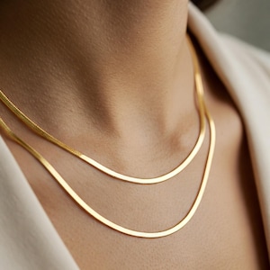 Könnte beinhalten: Zwei goldfarbene, flache Schlangenkettchen, die auf dem Hals einer Person übereinander liegen. Die Halsketten sind dünn und haben eine glatte, reflektierende Oberfläche. Die Person trägt einen weißen Blazer.