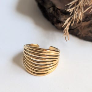 Könnte beinhalten: Ein goldfarbener Ring mit einem mehrschichtigen Design. Der Ring hat mehrere horizontale Bänder, die ein strukturiertes, wellenartiges Aussehen erzeugen. Der Ring befindet sich vor einem neutralen Hintergrund mit natürlichen Elementen.