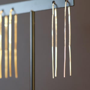 Könnte beinhalten: Goldfarbene Ohrhänger an einem schwarzen Ständer. Die Ohrringe haben ein zartes, lineares Design, einige mit einer kettenartigen Textur. Die Ohrringe sind vor einem neutralen Hintergrund aufgehängt.