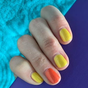 May include: Close-up of a hand with painted nails. The nails are painted in a mix of yellow and orange colors. The hand is resting on a purple surface, with a turquoise background.