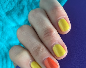May include: Close-up of a hand with painted nails. The nails are painted in a mix of yellow and orange colors. The hand is resting on a purple surface, with a turquoise background.