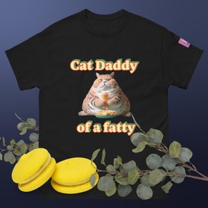 Może przedstawiać: Czarna koszulka z napisem "Cat Daddy of a fatty" w kolorze pomarańczowym i zielonym. Koszulka przedstawia kreskówkowego kota trzymającego szklankę piwa i talerz z jedzeniem. Dwa żółte makaroniki na pierwszym planie.