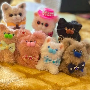 May include: A collection of handmade plush cat figures in various colors, including tan, pink, white, and black. Each cat has unique features like bow ties, hats, and decorative accents. The plush toys are arranged on a golden surface.