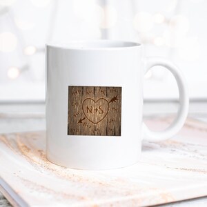 May include: White ceramic mug with a brown wood-grain design featuring a heart with the initials "N+S" carved into it. The heart is surrounded by arrows pointing in different directions.