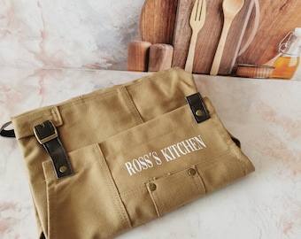 May include: A tan canvas apron with brown leather accents and the text "ROSS'S KITCHEN" in white. The apron has pockets and a buckle closure. Wooden cutting boards, a fork, and a spoon are in the background.