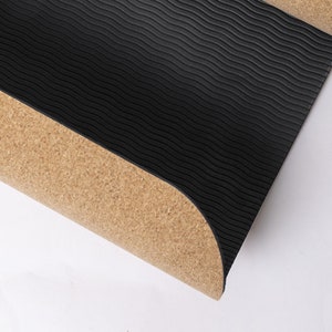 May include: A black yoga mat with a textured, wave-like pattern on the top surface. The bottom layer is a natural cork material. The mat is partially rolled, revealing the cork underside. The yoga mat is designed for exercise and fitness.