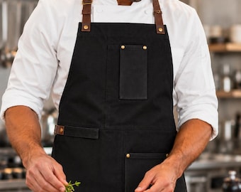 May include: A black apron featuring brown leather straps with brass-colored metal hardware, including buckles and rivets. The apron includes a vertical chest pocket and a lower pocket, both secured with brass-colored rivets at the corners.