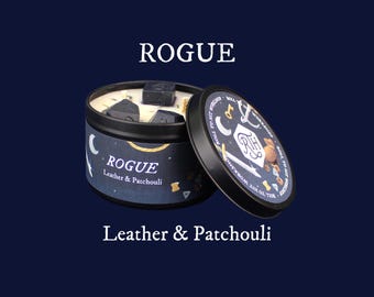 May include: A black metal candle tin with the word "ROGUE" in white, and "Leather & Patchouli" below. The lid is off, revealing the candle and black stones. The lid has a design with text and illustrations. The background is dark blue.