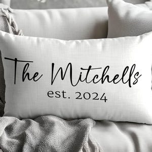 May include: A rectangular white throw pillow featuring black text that reads The Mitchells in a cursive script font, followed by the text est. 2024 in a simple sans-serif font centred below the family name on the front of the pillow cover.
