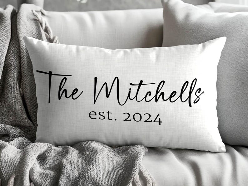 May include: A rectangular white throw pillow featuring black text that reads The Mitchells in a cursive script font, followed by the text est. 2024 in a simple sans-serif font centred below the family name on the front of the pillow cover.